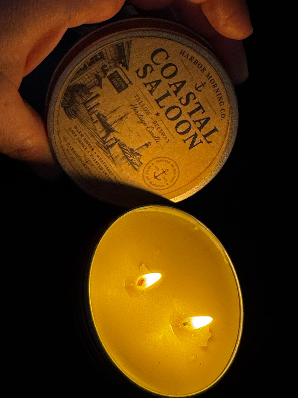 Candle inside a container labeled 'Coastal Saloon' held by a hand against a dark background.