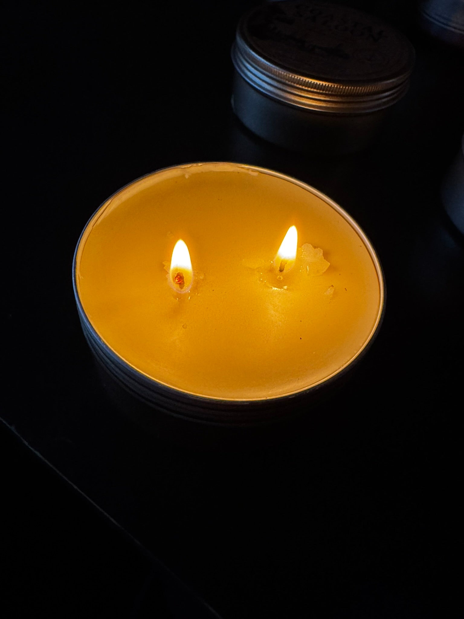 Two lit candles in a dark setting with a metallic container in the background.
