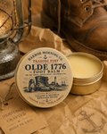 Oldé 1776 foot balm tin with a rustic background featuring boots and lantern.