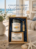 Harbor Morning Co. Coastal Welcome Box open on a sun-bleached wooden table in a coastal home — featuring a hand-poured candle, goat milk soap, and teal matches in kraft box with navy crinkle padding, blue seas visible through large windows