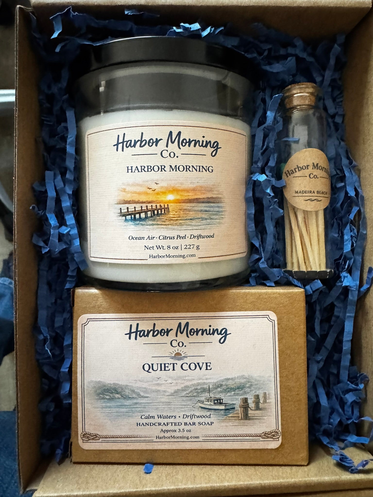 Harbor Morning Co. Coastal Welcome Box open product detail — hand-poured candle, goat milk soap, and teal matches in kraft box with navy blue crinkle padding