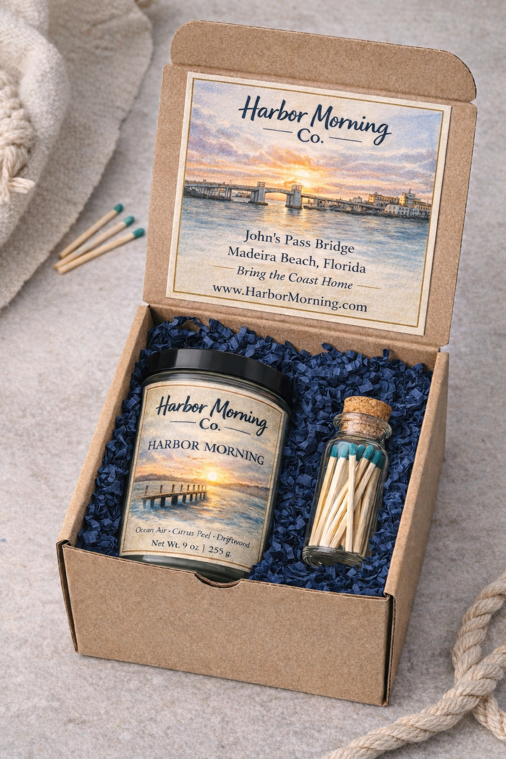 Harbor Morning Co. Founder Batch Gift Box with postcard, candle, and matches in kraft box with navy blue crinkle padding, sea rope, and towel nearby