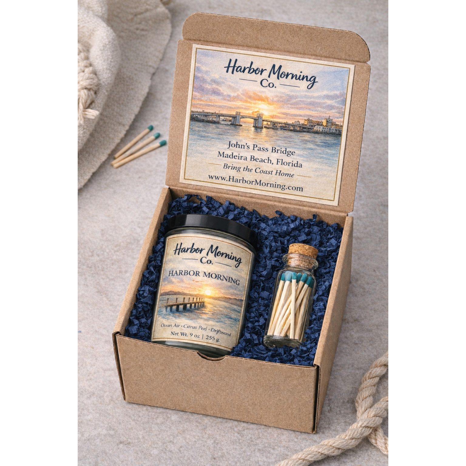 Harbor Morning Co. Founder Batch Gift Box with postcard, candle, and matches in kraft box with navy blue crinkle padding, sea rope, and towel nearby